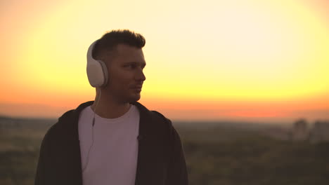 Smiling-and-laughing-handsome-young-man-listening-music-from-his-smartphone-in-wireless-headphones.-In-slow-motion-a-man-stands-on-the-roof-at-sunset-and-looks-at-the-beautiful-view-from-the-height-of-the-city